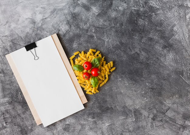 Fusilli crudo con tomates y hojas de albahaca con papel en blanco en el portapapeles con fondo texturado