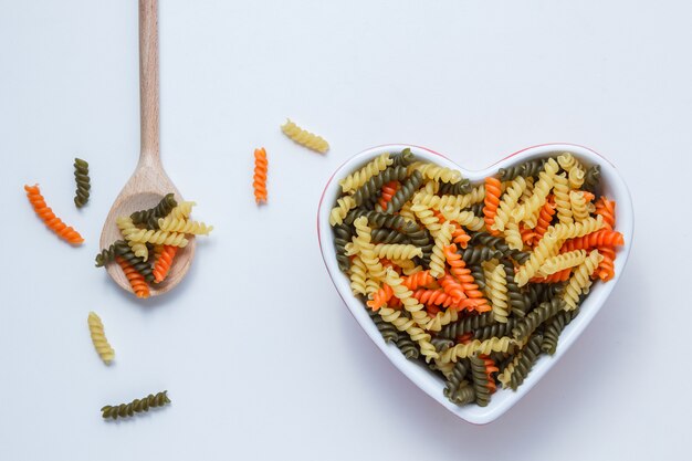 Fusilli pasta en tazón y cuchara de madera en mesa blanca, vista superior.