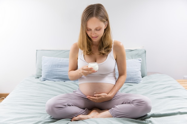 Futura madre pensativa comiendo yogurt en el dormitorio