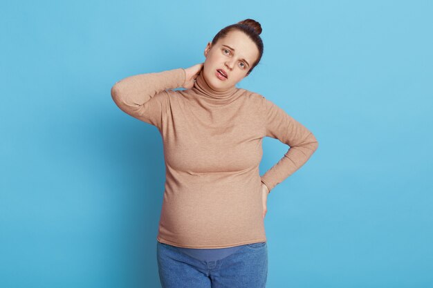 Futura mamá con dolor de cuello posando aislada sobre pared azul, manteniendo la mano en la cadera, con expresión triste, vestida con un suéter beige y jeans, muy cansada de la barriga embarazada.