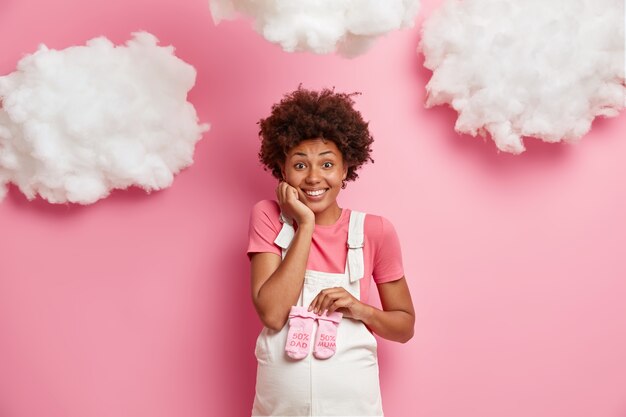 La futura mamá feliz espera un bebé, sostiene los calcetines rosados del niño sobre la barriga, se viste con un overol de mezclilla, tiene una expresión alegre, posa contra la pared rosada, elige la ropa del bebé. Concepto de embarazo