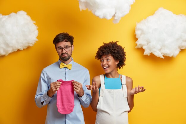 El futuro papá disgustado sostiene la ropa del niño, triste por saber el género del bebé, quiere tener un hijo, la esposa embarazada está cerca. Los futuros padres casados esperan recién nacido, eligen cosas para el niño