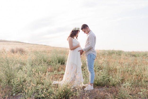 Los futuros padres están parados en un cálido día soleado en el campo, vestidos con ropa casual.