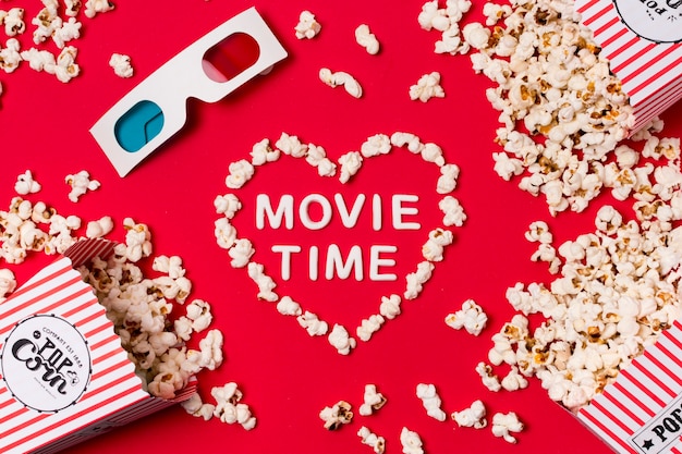 Gafas 3d; Las palomitas de maíz se derramaron de la caja con el texto de la hora de la película en forma de corazón sobre fondo rojo