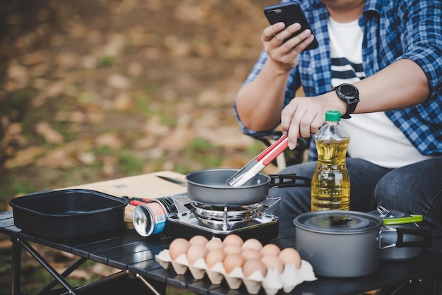 Gafas de hombre viajero asiático freír un sabroso huevo frito en una sartén caliente en el camping Cocina al aire libre viajar camping estilo de vida concepto