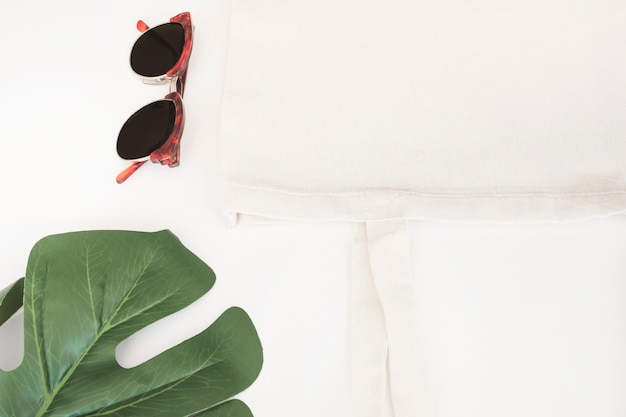 Gafas de sol, bolso de tela blanca y hoja de monstera sobre fondo blanco