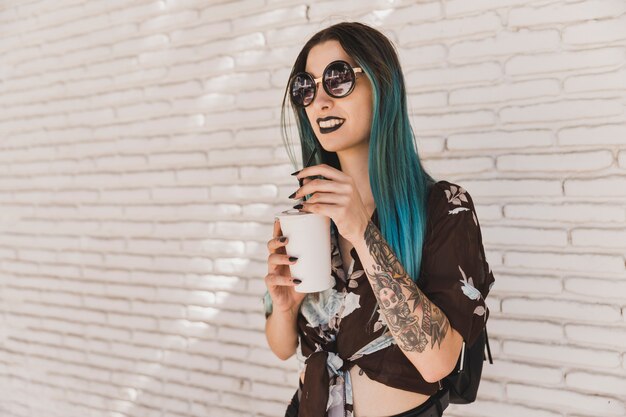 Gafas de sol que llevan hermosas modernas de la mujer joven que sostienen la taza de café disponible