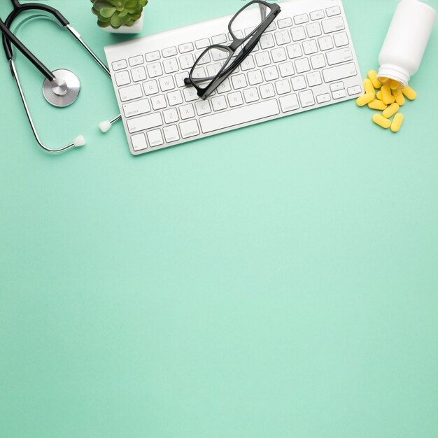 Las gafas en el teclado inalámbrico cerca de las píldoras derramaron la botella frontal y el estetoscopio sobre la superficie verde