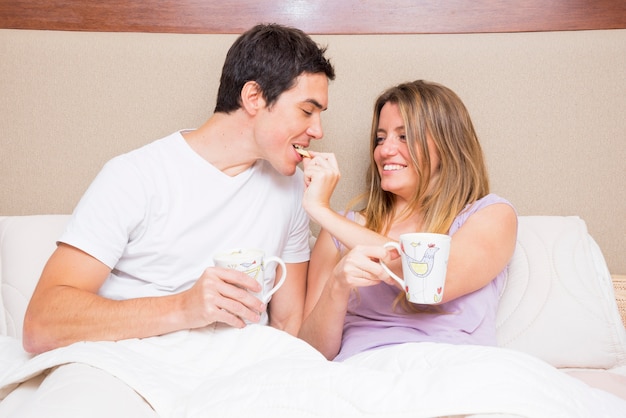 Galleta de alimentación de la mujer feliz a su novio que se sienta en cama