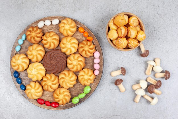 Galletas en una bandeja de madera rodeada de caramelos y en un recipiente con un paquete disperso de setas de chocolate sobre fondo de mármol. Foto de alta calidad