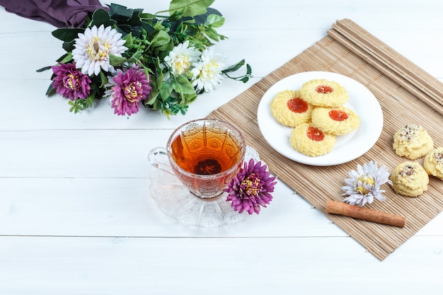 Galletas, canela en un mantel con flores, taza de té vista de ángulo alto sobre un fondo de tablero de madera blanca