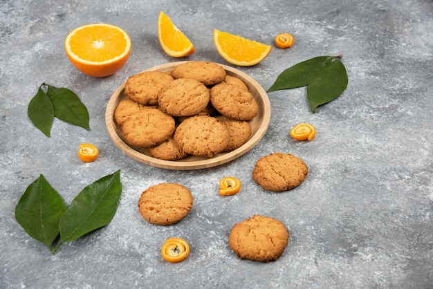 Galletas caseras sobre tabla de madera y rodajas de naranja con hojas sobre superficie gris.