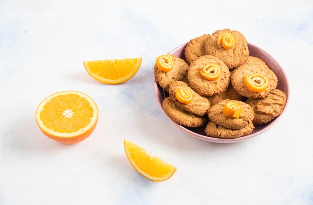 Galletas caseras en tazón de fuente rosa y rodajas de naranja sobre mesa blanca.