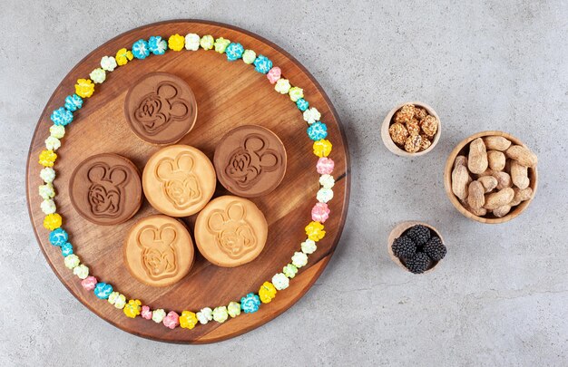 Galletas y un círculo de caramelos sobre una tabla de madera junto a tazones de maní y mullberries sobre fondo de mármol. Foto de alta calidad
