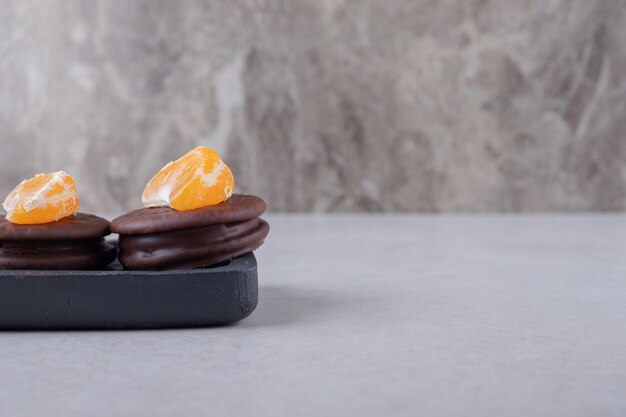 Galletas cubiertas de chocolate con rodaja de mandarina en la bandeja de madera sobre la mesa de mármol.