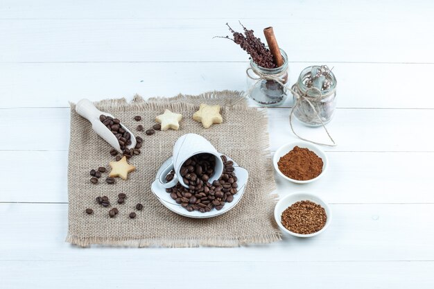 Galletas de estrellas, granos de café en un pedazo de saco con un tazón de café instantáneo, un tarro de hierbas de cerca sobre un fondo de tablero de madera blanca