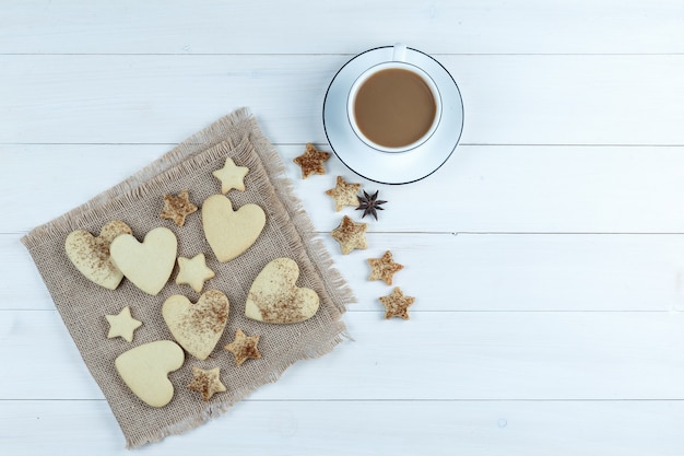 Galletas en forma de corazón y estrella en un pedazo de saco con galletas estrella, taza de café plana yacía sobre un fondo de tablero de madera blanca
