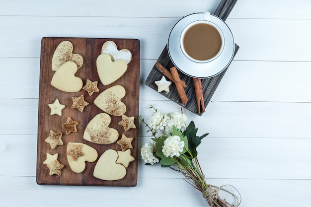 Galletas en forma de corazón y estrella sobre una tabla de cortar de madera con una taza de café, flores, canela plana yacía sobre un fondo de tablero de madera blanca