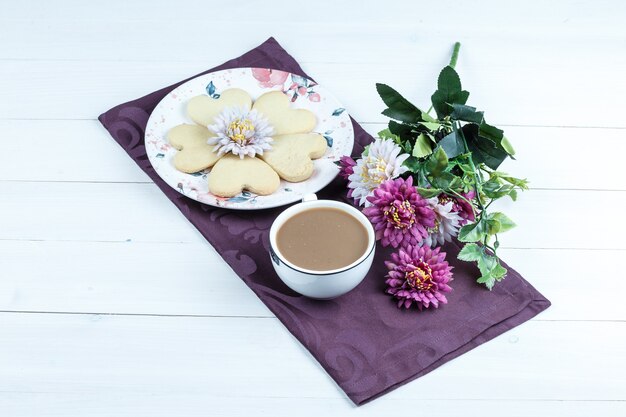 Galletas en forma de corazón, taza de café en un mantel púrpura con flores vista de ángulo alto sobre un fondo de tablero de madera blanca