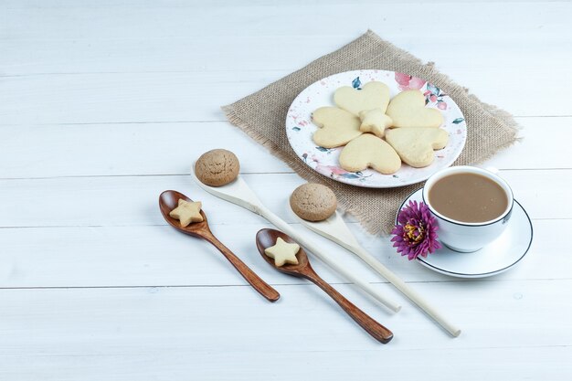 Galletas en forma de corazón de vista de ángulo alto en un pedazo de saco con flor, galletas en cucharas de madera, taza de café sobre fondo de tablero de madera blanca. horizontal