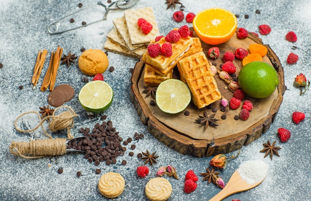 Galletas con harina, hierbas, frutas, especias, choco, colador plano sobre tablero de madera y fondo de estuco