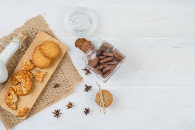 Galletas marrones de la vista superior en frasco de vidrio con una jarra de leche, galletas en la tabla de cortar y un pedazo de saco en la superficie blanca