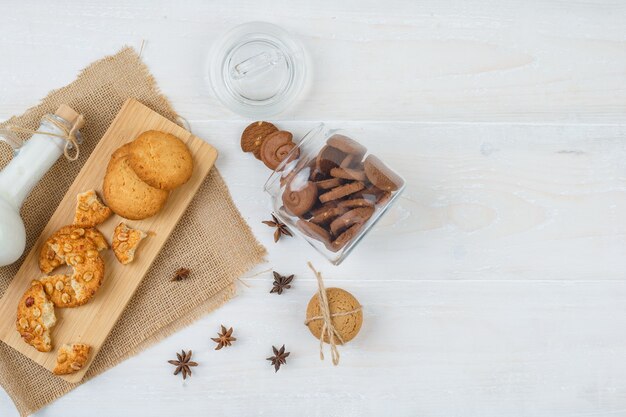 Galletas marrones de la vista superior en frasco de vidrio con una jarra de leche, galletas en la tabla de cortar y un pedazo de saco en la superficie blanca