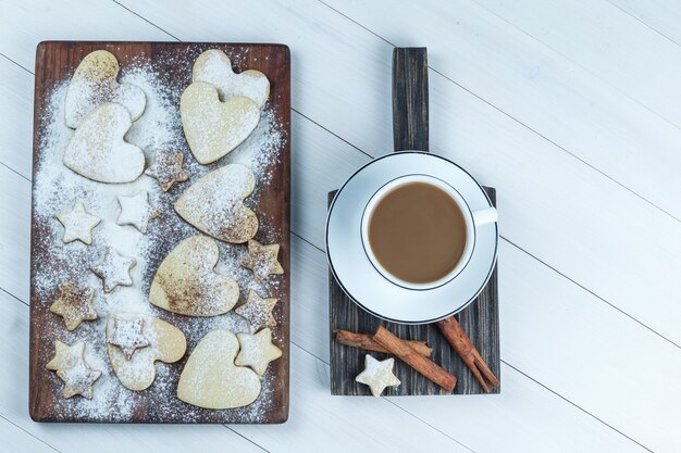 Galletas planas en forma de corazón y estrella sobre tabla de cortar de madera con taza de café, canela sobre fondo de tablero de madera blanca. horizontal