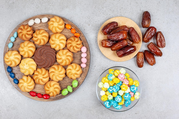 Galletas rodeadas de caramelos sobre tabla de madera junto al cuenco de caramelos y dátiles sobre superficie de mármol.