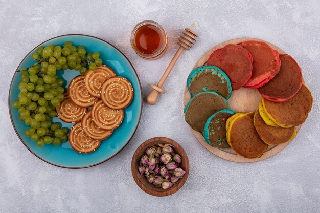 Galletas de vista superior con uvas verdes en una placa azul con cogollos secos de miel y panqueques multicolores en un soporte sobre un fondo blanco.