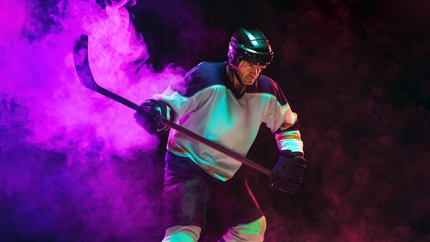 Ganador. Jugador de hockey masculino con el palo en la cancha de hielo y una pared de color neón oscuro. Deportista con equipo, casco practicando. Concepto de deporte, estilo de vida saludable, movimiento, bienestar, acción.