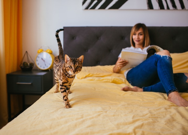 Gato de Bengala se acerca a una mujer mientras lee en la cama