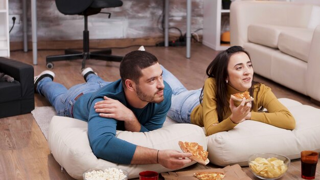 Gato durmiendo mientras sus dueños ven la televisión y comen pizza. Pareja sentada en el suelo.