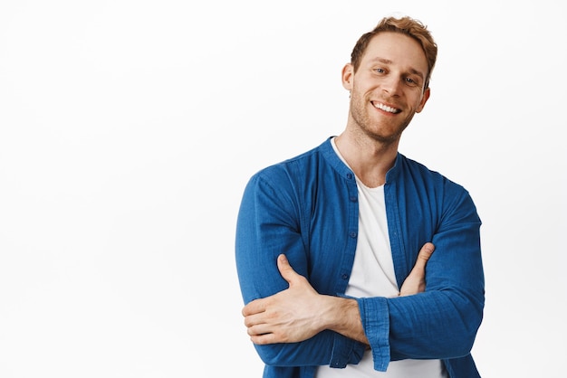 Genial y apuesto hombre pelirrojo sonriendo con confianza cruzando los brazos en el pecho y luciendo feliz sin molestias de pie en una pose relajada casual contra el fondo blanco