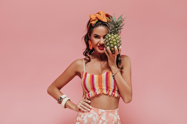 Genial chica maravillosa con accesorios modernos de cabello oscuro y manicura blanca en traje brillante cubre su rostro con piña y sonrisas