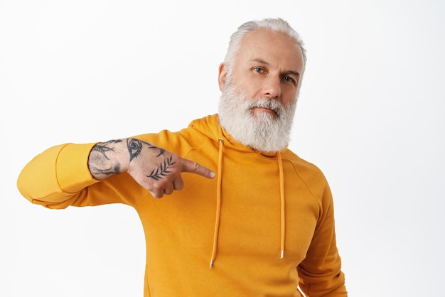 Foto gratuita genial hombre mayor con tatuajes apuntándose a sí mismo abuelo barbudo con capucha naranja que se autopromociona alardeando de su logro de pie sobre fondo blanco