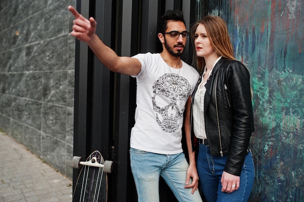 Genial pareja multirracial posando contra la pared con un novio árabe de longboard mostrando algo a su chica europea