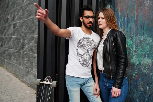 Genial pareja multirracial posando contra la pared con un novio árabe de longboard mostrando algo a su chica europea