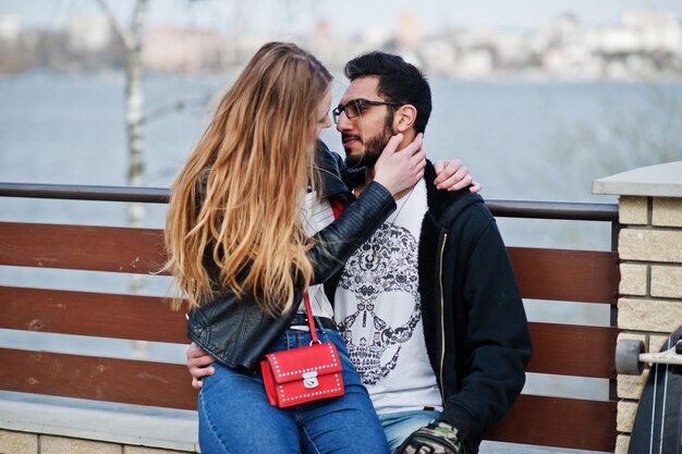 Genial pareja multirracial sentada contra el fondo del lago y abrazos juntos