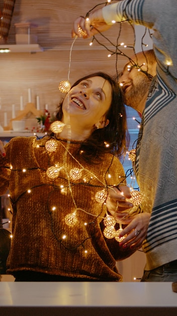 Gente alegre que se enreda en luces festivas mientras decoran el hogar para la celebración de la víspera de Navidad. Hombre y mujer sonriendo, tratando de desenredar la guirnalda de bombillas de luces centelleantes.