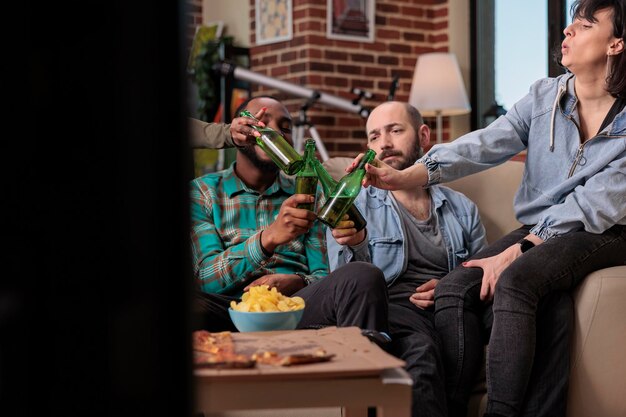 Gente alegre tintineando botellas de cerveza y brindando celebración, haciendo gestos de alegría con bebidas en la reunión de amistad. Amigos brindando con bebida alcohólica, evento social en casa.