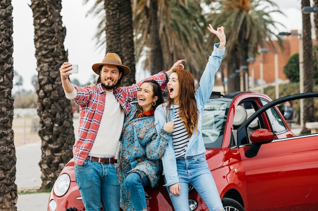 Foto gratuita gente alegre tomando selfie cerca de auto rojo en la calle