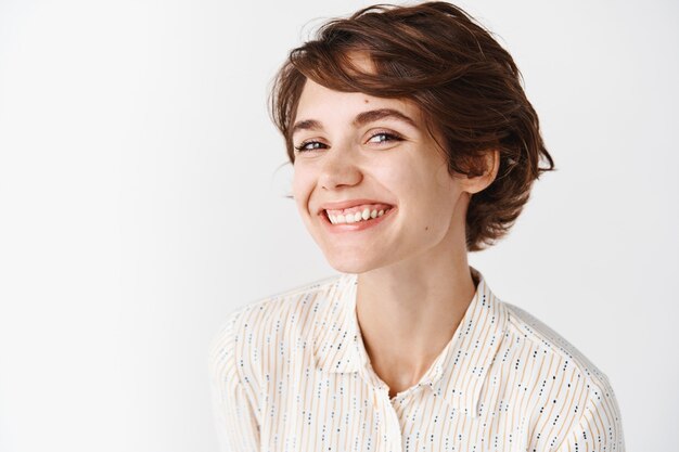 Gente auténtica y sincera. Hermosa mujer joven en blusa con peinado corto, sonriendo feliz, de pie en la pared blanca