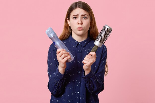 Gente, belleza, concepto de aseo. La joven disgustada pone mala cara con los labios vestidos con una camisa elegante, posa sobre la pared rosada, sostiene la laca y el peine, acude al peluquero para hacer el peinado.