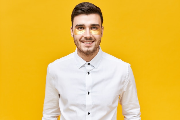 Gente, belleza y estilo de vida. Hombre de negocios exitoso con camisa blanca haciendo rutina matutina, posando aislado en parches debajo de los ojos para reducir la hinchazón antes de una reunión de negocios importante