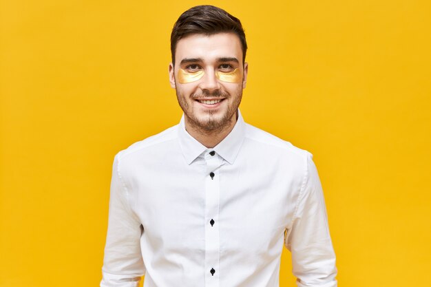 Gente, belleza y estilo de vida. Hombre de negocios exitoso con camisa blanca haciendo rutina matutina, posando aislado en parches debajo de los ojos para reducir la hinchazón antes de una reunión de negocios importante