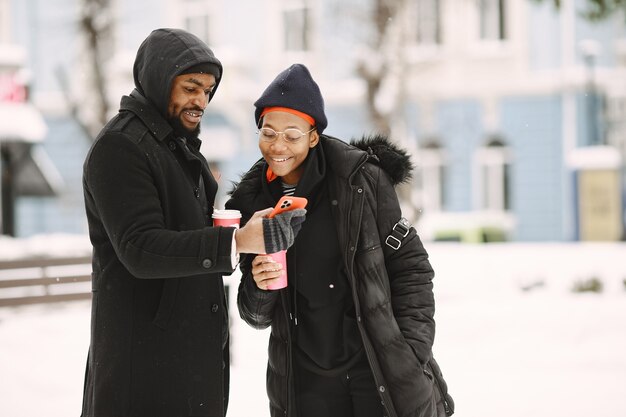 La gente camina afuera. Día de invierno. Pareja africana con café.