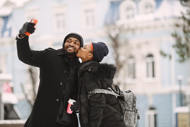 La gente camina afuera. Día de invierno. Pareja africana con café.