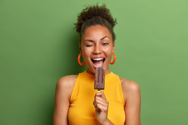 Foto gratuita gente comida chatarra poco saludable y concepto de horario de verano. alegre mujer de piel oscura guiña el ojo come un delicioso helado de chocolate se divierte viste ropa casual brillante aislada sobre la pared verde