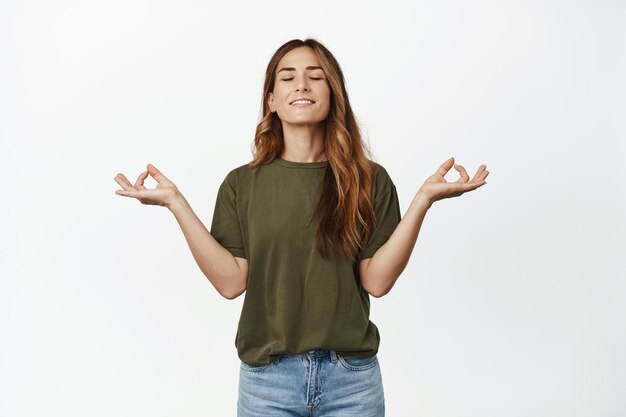 Gente consciente. Mujer tranquila y relajada de mediana edad que respira en paz, inhala aire, medita con los ojos cerrados y una sonrisa complacida, practica yoga, se enfrenta a un fondo blanco